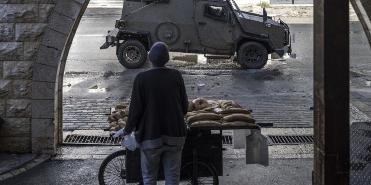 A Palestinian bread seller stands by his cart while an Israeli army vehicle drives by at the entrance of the Jenin refugee camp, in Jenin, on December 13, 2023 amid ongoing battles between Israel and the Palestinian movement Hamas. Israeli forces killed four Palestinians in Jenin on December 12, the Palestinian health ministry reported without further details on the fatalities, while the Palestinian Red Crescent Society said they were killed in a drone strike in the old town. (Photo by MARCO LONGARI / AFP)